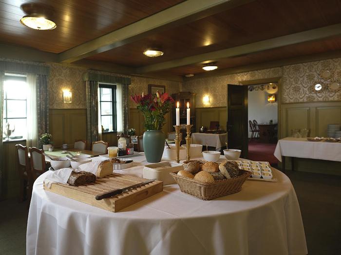 Et rundt bord med brød, rundstykker, smør og stearinlys i en hyggelig spisestue med træloft, mønstret tapet og blødt, naturligt lys.