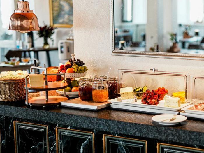 En morgenmadsbuffet med syltetøjsglas, ost, frisk frugt, grøntsager, kager og brød på en disk med et spejl og en spiseplads i baggrunden.