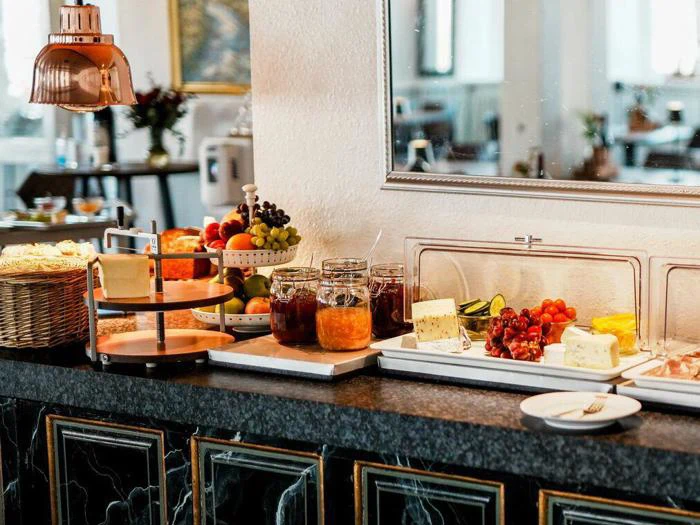 Morgenmadsbuffet med forskellige oste, syltetøj, frisk frugt, grøntsager, brød og kager på en marmordisk i et godt oplyst spiseområde.
