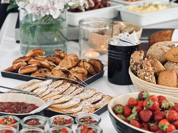 En brunchbuffet med pandekager, croissanter, diverse rundstykker, jordbær og syltetøjsglas på en hvid dug. Blomster og et tændt stearinlys i baggrunden.