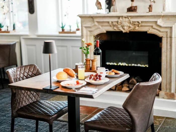 Et hyggeligt restaurantbord dækket til to med morgenmad, brød, vindruer, juice, kaffe og en flaske vin nær en dekorativ pejs.