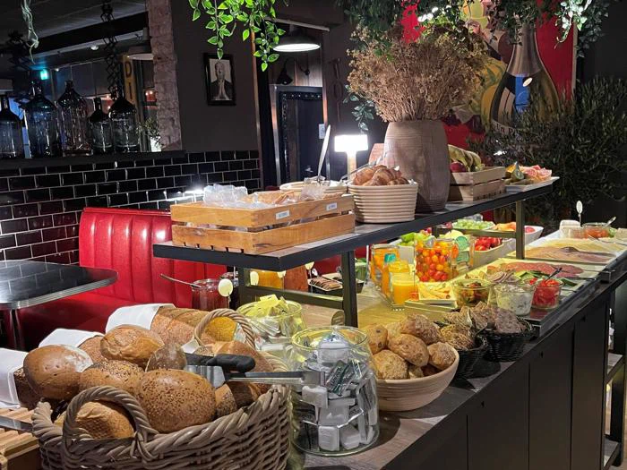 Et buffetbord i en restaurant viser brød, kager, frugt, ost, pålæg, juice og grønt, med hyggelige siddepladser og varm belysning i baggrunden.