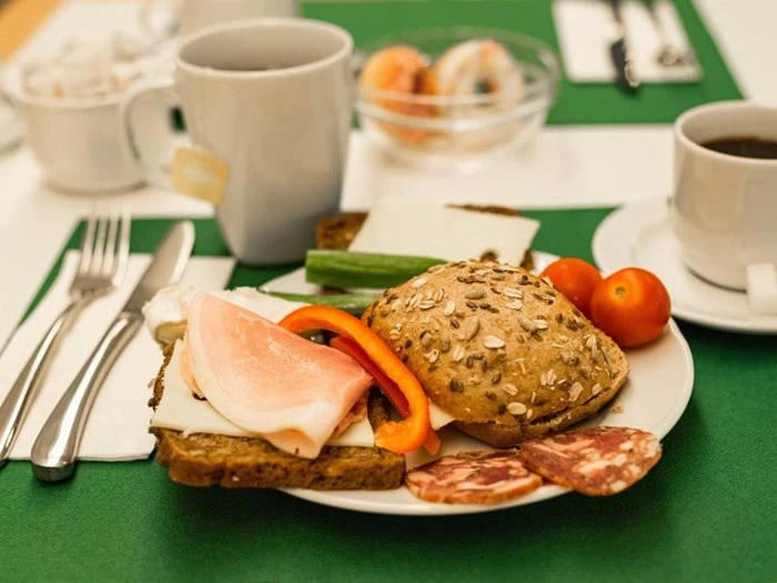 En morgenmadstallerken med brød med kerner, ost i skiver, skinke, salami, en skive rød peberfrugt, agurk og cherrytomater. Kaffe og redskaber står på bordet.