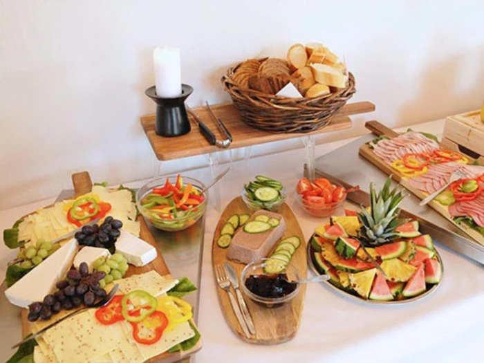 Et buffetbord med forskellige oste, vindruer, kød i skiver, friske grøntsager, brød i en kurv, vandmelonskiver og et stearinlys i en holder.