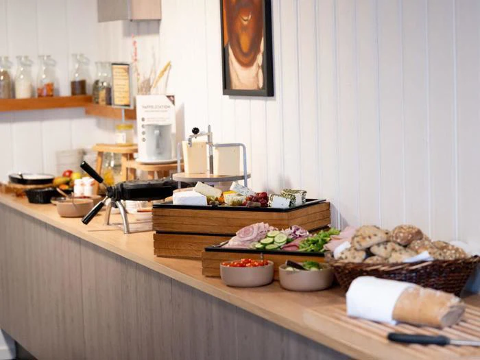 En morgenmadsbuffet med forskellige oste, pålæg, brød, friske grøntsager og smørepålæg arrangeret i skåle og kurve på en trædisk.