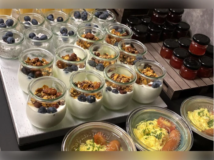 En udstilling af glas fyldt med yoghurt, granola og blåbær ved siden af glas med rød marmelade og beholdere med en gul skål med krydderurter.