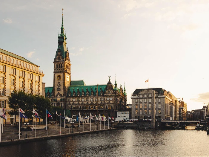 Udsigt over Hamborgs rådhus og nærliggende bygninger ved solnedgang, med flag langs havnefronten og deres refleksioner synlige i vandet.