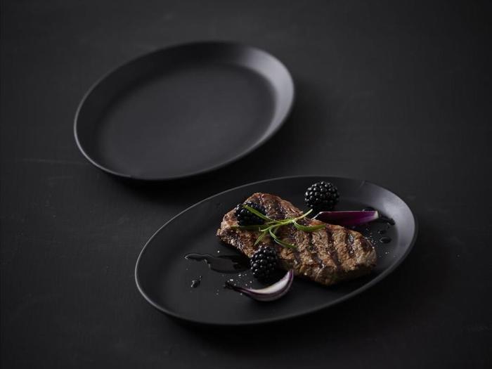 En grillet bøf garneret med brombær, rødløgsskiver og krydderurter på en sort tallerken med en tom sort tallerken i baggrunden.