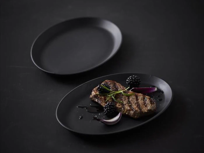 En grillet bøf garneret med brombær, rødløgsskiver og krydderurter på en sort tallerken med en tom sort tallerken i baggrunden.