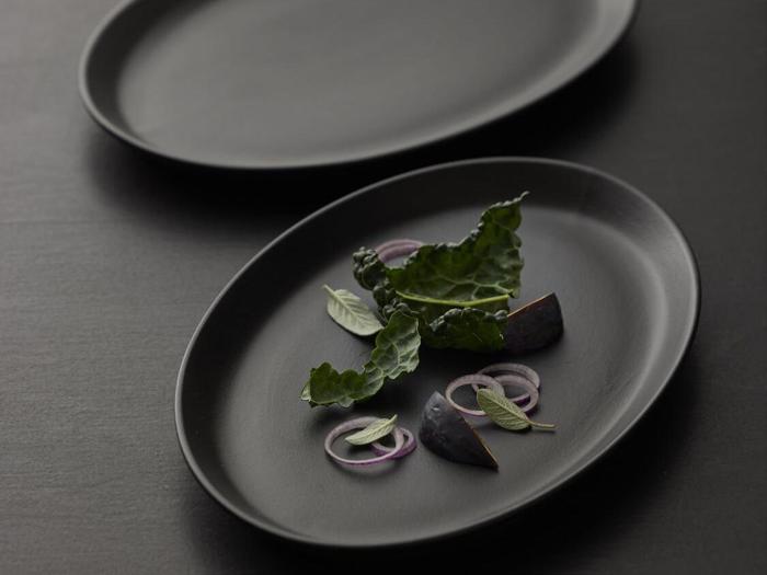 En sort oval tallerken med bladgrønt, rødløgsringe, basilikumblade og to figenkiler, kunstfærdigt arrangeret på en mørk overflade.