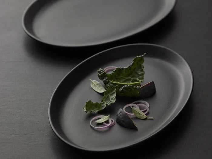 En sort oval tallerken med bladgrønt, rødløgsringe, basilikumblade og to figenkiler, kunstfærdigt arrangeret på en mørk overflade.