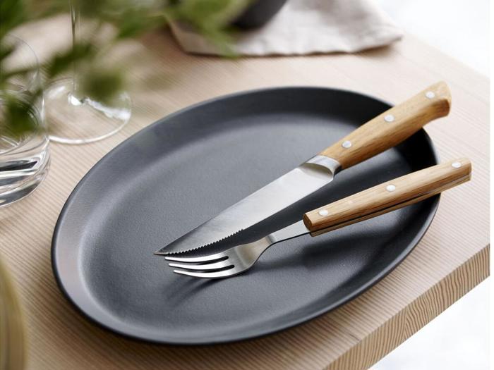 En sort oval tallerken med en gaffel og en savtakket kniv, begge med træhåndtag, placeret på et lyst træbord.