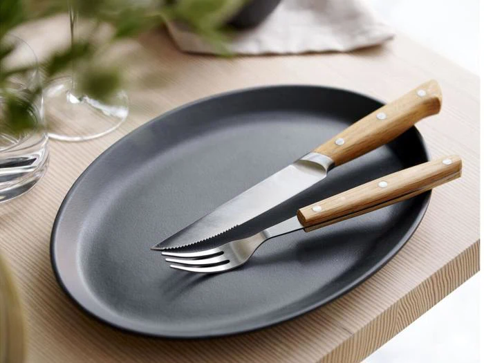 En sort oval tallerken med en gaffel og en savtakket kniv, begge med træhåndtag, placeret på et lyst træbord.
