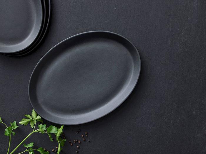En sort oval tallerken på en mørk overflade med persilleblade og sorte peberkorn i nærheden; stablede tallerkener er delvist synlige i hjørnet.