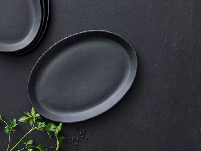 En sort oval tallerken på en mørk overflade med persilleblade og sorte peberkorn i nærheden; stablede tallerkener er delvist synlige i hjørnet.