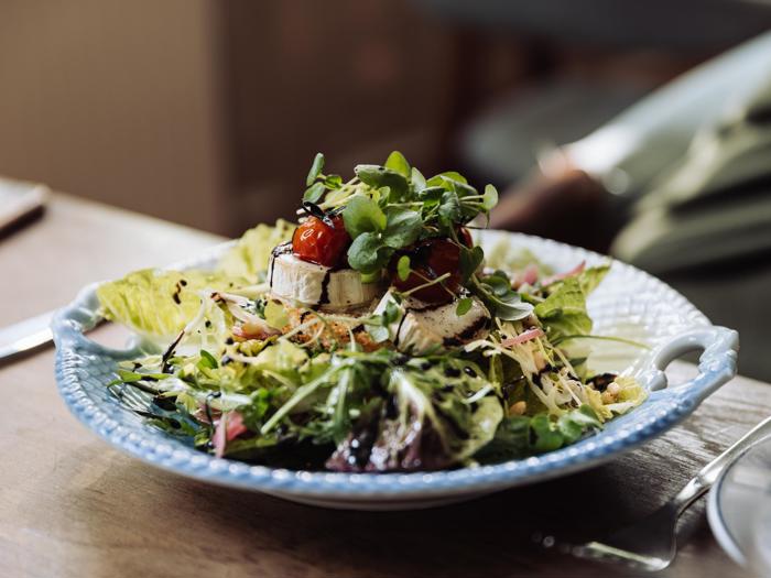 En farverig salat med blandet grønt, gedeost, cherrytomater og balsamico-glasur på en blå tallerken, placeret på et træbord.