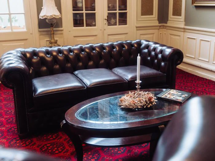 En mørkebrun Chesterfield-sofa i læder står bag et sofabord i glas med et stearinlys, en dekorativ bordskåner og magasiner på et rødmønstret tæppe i et elegant rum.