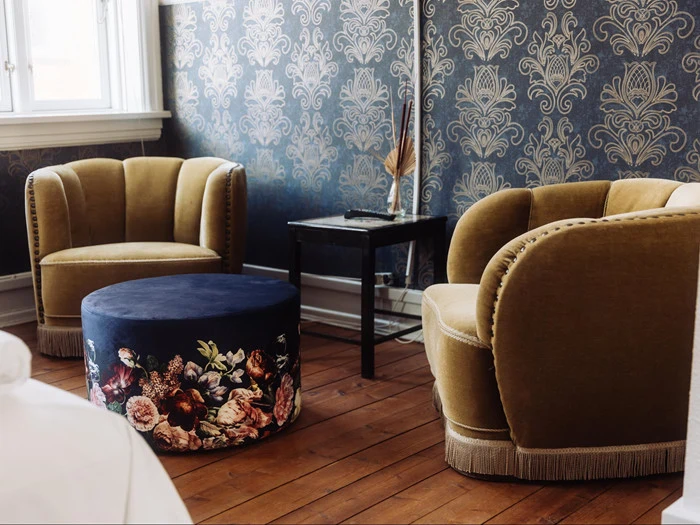 To gule fløjlslænestole og en blomstermønstret rund ottoman står på et trægulv mod et blåblomstret tapet i et hyggeligt rum i vintagestil.