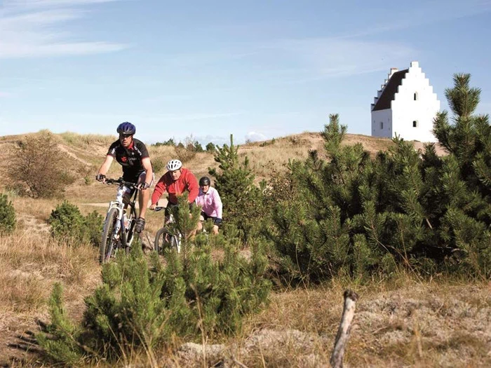 Tre personer kører på mountainbike på en græsklædt sti omgivet af små fyrretræer med en hvid kirkelignende bygning i baggrunden under en klar himmel.