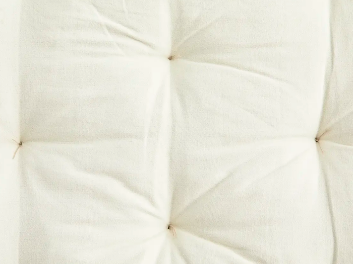 Nærbillede af en cremefarvet, tuftet stofoverflade med synlige syede knapper, der skaber en blød, quiltet tekstur.