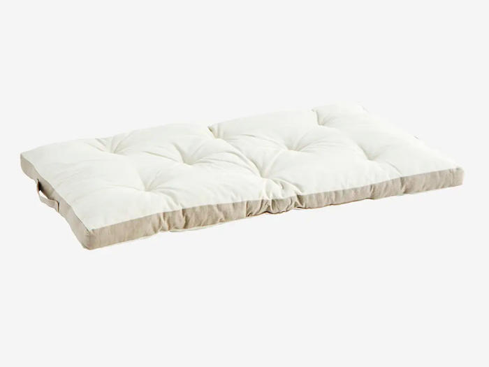 En rektangulær, hvid, tuftet gulvpude med beige kanter og et stofhåndtag på den ene side, placeret på en almindelig hvid baggrund.