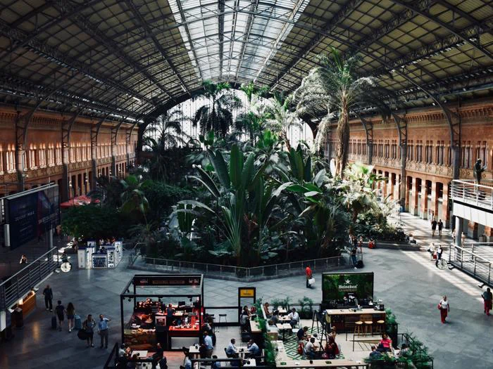 En stor indendørs togstation med højt glasloft, frodige tropiske planter i midten og mennesker, der går og sidder på caféer og bænke.