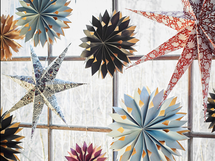 Papirstjerner og snefnugpynt i forskellige farver og mønstre hænger foran et vindue med nøgne træer udenfor.
