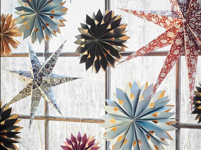 Papirstjerner og snefnugpynt i forskellige farver og mønstre hænger foran et vindue med nøgne træer udenfor.