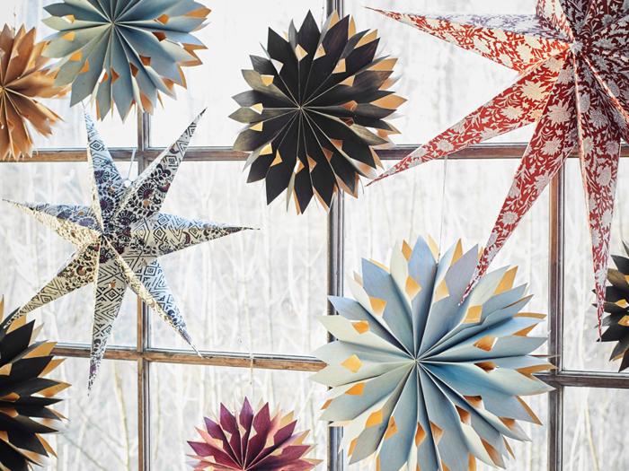 Farverige papirstjerner og snefnugdekorationer hænger foran et vindue med en snedækket eller frostklar udendørs baggrund, der er synlig gennem glasset.
