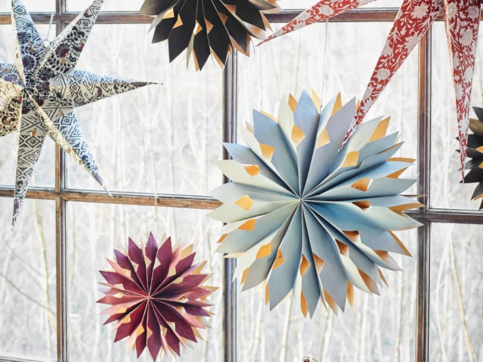 Stjernedekorationer af papir i forskellige farver og mønstre hænger foran et vindue med en vinterlig, bladløs skov udenfor.