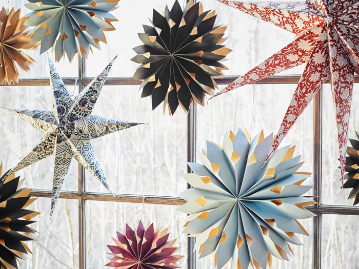 Farverige stjerne- og blomsterdekorationer af papir hænger foran et vindue og lader lyset skinne gennem de udskårne mønstre.