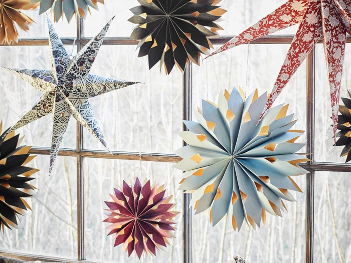 Farverige papirstjerner og blomsterdekorationer hænger foran et vindue med en sløret udendørs baggrund.