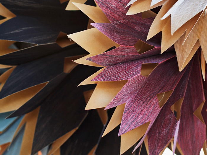 Nærbillede af lagdelte, spidse papirformer i brune, sorte og rødlilla nuancer, der ligner abstrakte blade eller kronblade, som overlapper hinanden.
