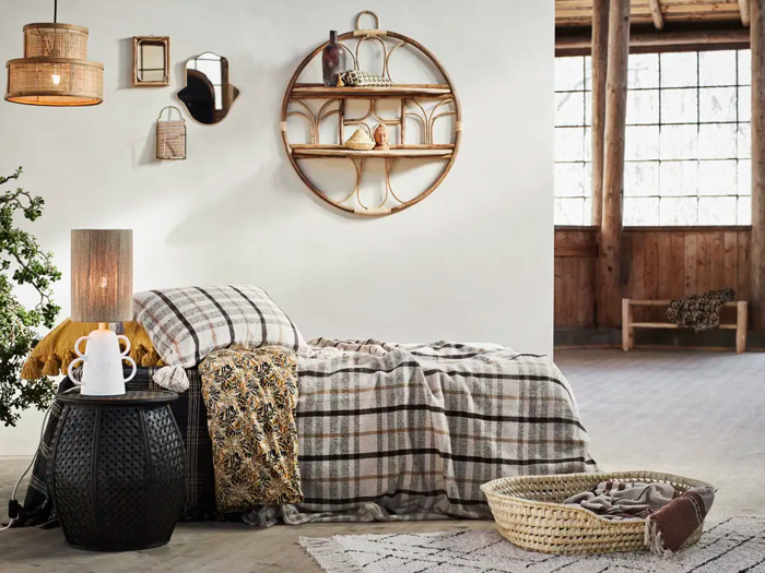 Hyggeligt soveværelse med ternet sengetøj, pyntepuder, et natbord med lampe og tekande, væghylder og en vævet kæledyrsseng på et mønstret tæppe nær store vinduer.