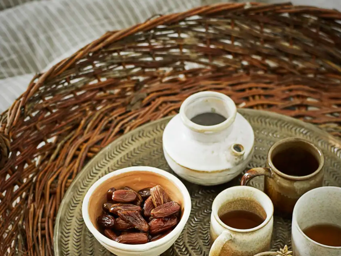 På en flettet bakke står en keramiktallerken med dadler, en lille hvid tekande og to brune kopper fyldt med te.