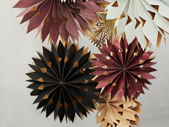 Papirstjernedekorationer i nuancer af sort, bordeaux, creme og beige er arrangeret på en hvid struktureret væg. Hver stjerne har spidse, lagdelte kronblade med udskårne detaljer.