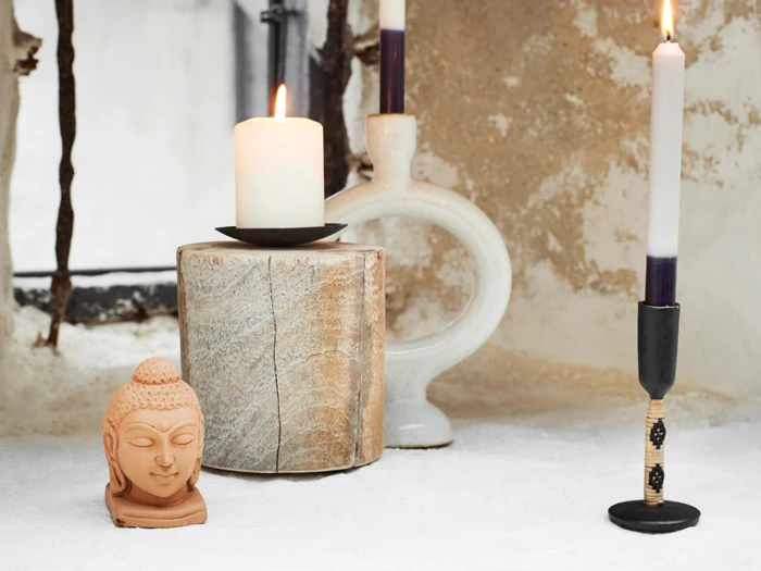 En lille buddhastatue, to tændte stearinlys i holdere og en træklods er anbragt på en hvid flade nær et rustikt vindue.