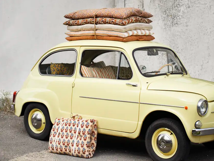 En lille gul veteranbil er parkeret ved en mur med flere mønstrede puder bundet på taget og en matchende taske placeret på jorden ved siden af.
