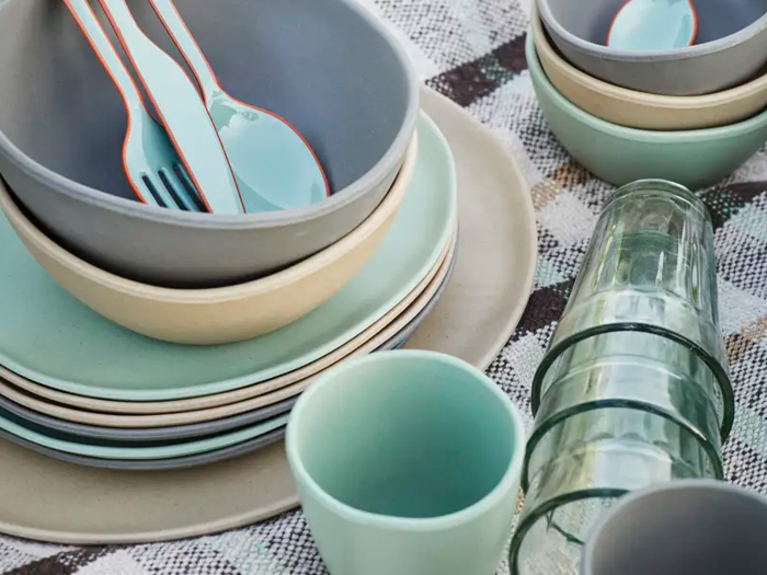 En stak pastelfarvede tallerkener, skåle, kopper og glasbeholdere på en ternet dug med lyseblå redskaber i en skål.