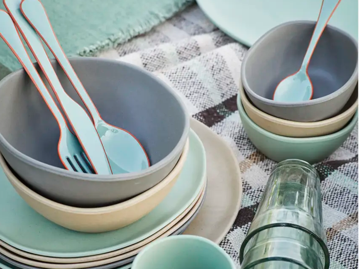 Stablede tallerkener, skåle og kopper i dæmpede farver med noget blåt bestik placeret i skåle, alt sammen arrangeret på et ternet klæde. Et grønt glas ligger på siden.