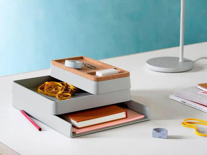 En moderne skrivebordsorganizer rummer papirvarer, notesbøger, elastikker og små genstande på et hvidt skrivebord ved siden af en lampe og bøger mod en blå væg.