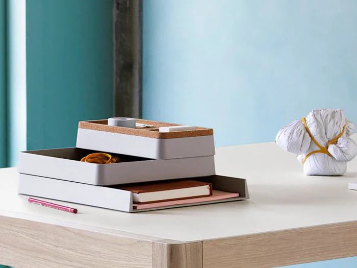 Et ryddeligt skrivebord med stablede grå og kork-organisatorer, en notesbog, blyant og elastik ved siden af et indpakket bundt bundet med snor.