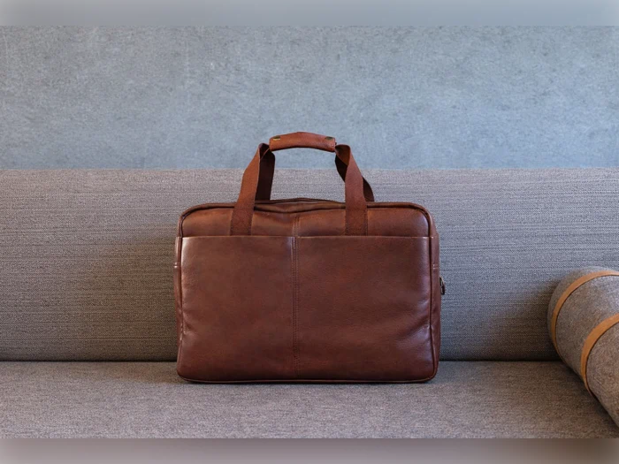 En brun lædermappe med håndtag ligger på en grå stofsofa med et sammenrullet tæppe ved siden af.