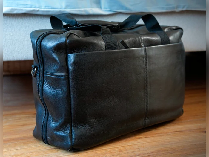 En sort lædertaske med håndtag står på et trægulv foran en beige sofa eller seng.