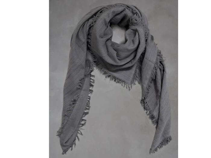 Et stort, gråt tørklæde med frynsede kanter er draperet mod en almindelig, grå baggrund.