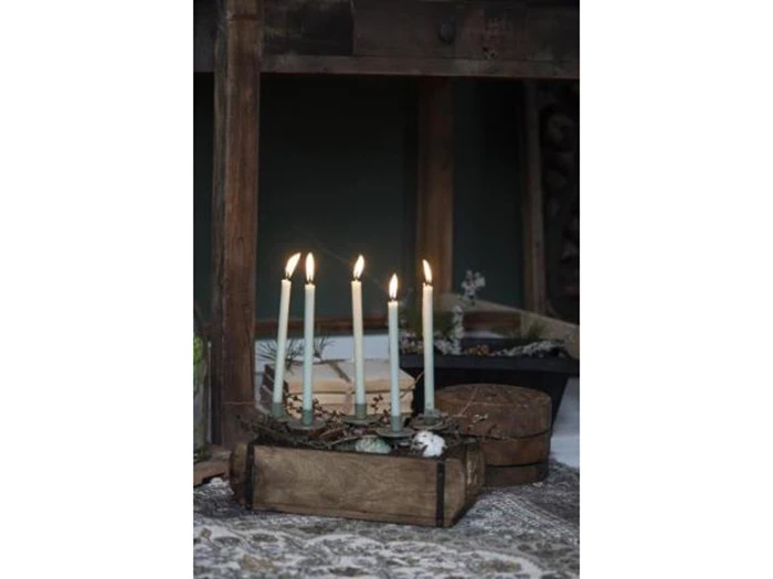 Fire tændte hvide stearinlys står oprejst i en rustik trækasse på et mønstret tæppe med et mørkt træbord og anden indretning i baggrunden.