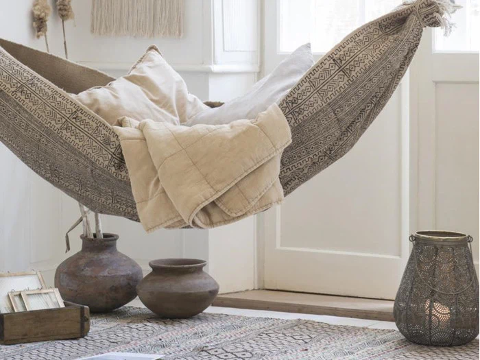 En mønstret hængekøje med beige puder og et tæppe hænger indendørs over et tæppe ved siden af lerkrukker, en lanterne og en stak bøger i et hyggeligt, lyst rum.