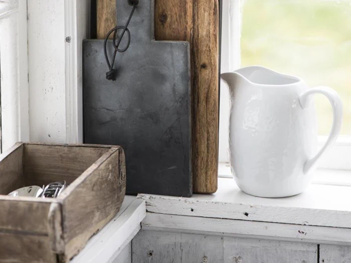 En hvid keramikkande står i vindueskarmen ved siden af en trækasse og to skærebrætter, et i træ og et i sort, i en rustik køkkenindretning.