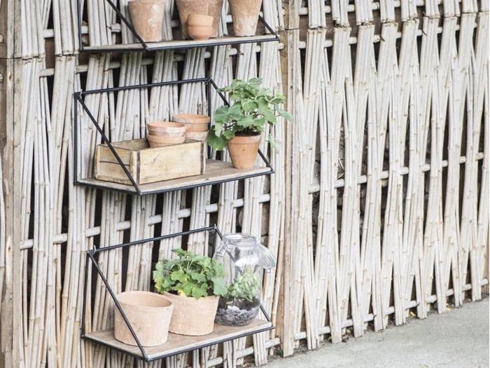 Tre vægmonterede hylder af metal og træ rummer potteplanter og en glaskrukke, der er fastgjort til et lyst, flettet hegn udendørs.