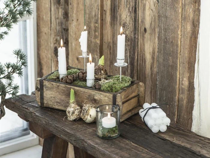 En rustik trækasse med hvide lys, mos, løg og kogler på en træbænk; ekstra lys og en glaslysestage står ved siden af.